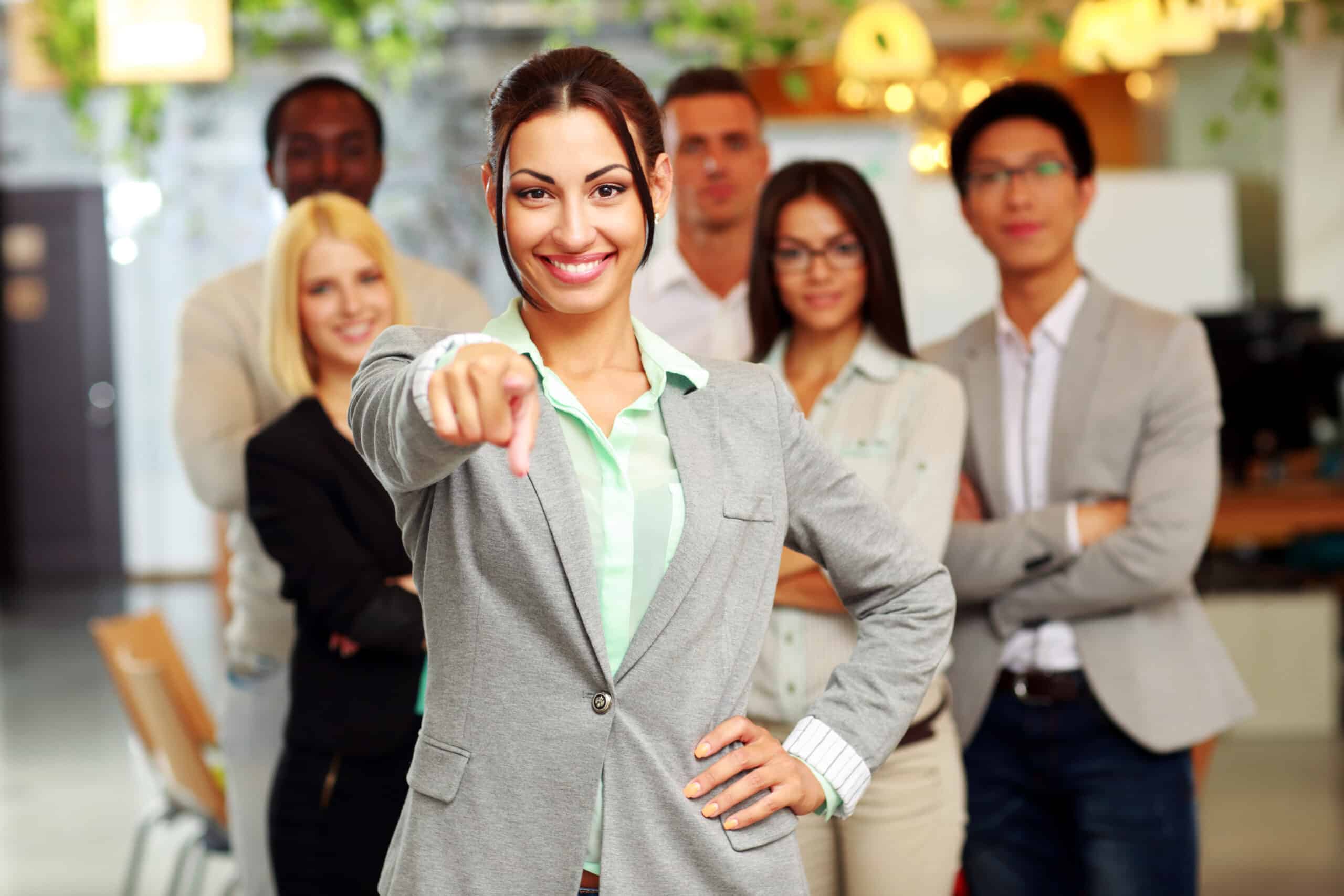 Smiling handsome businesswoman with team in office pointing finger at you scaled