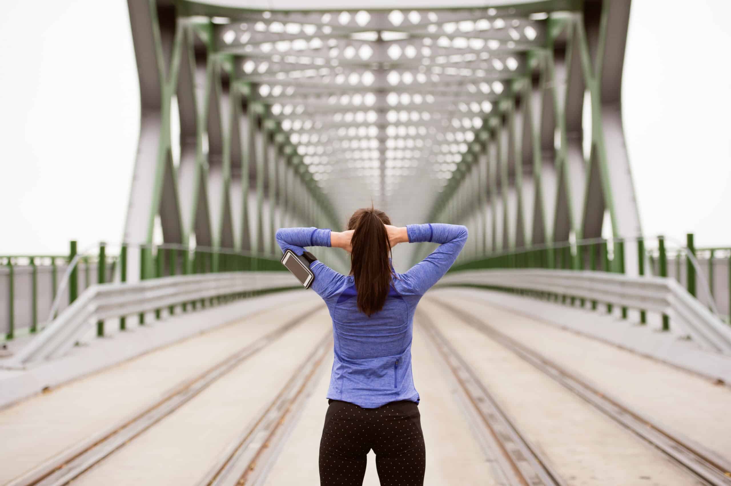 graphicstock beautiful young runner with smart phone in the city tracking her progress warming up and stretching on green steel bridge rear view rOln7D3Bz scaled